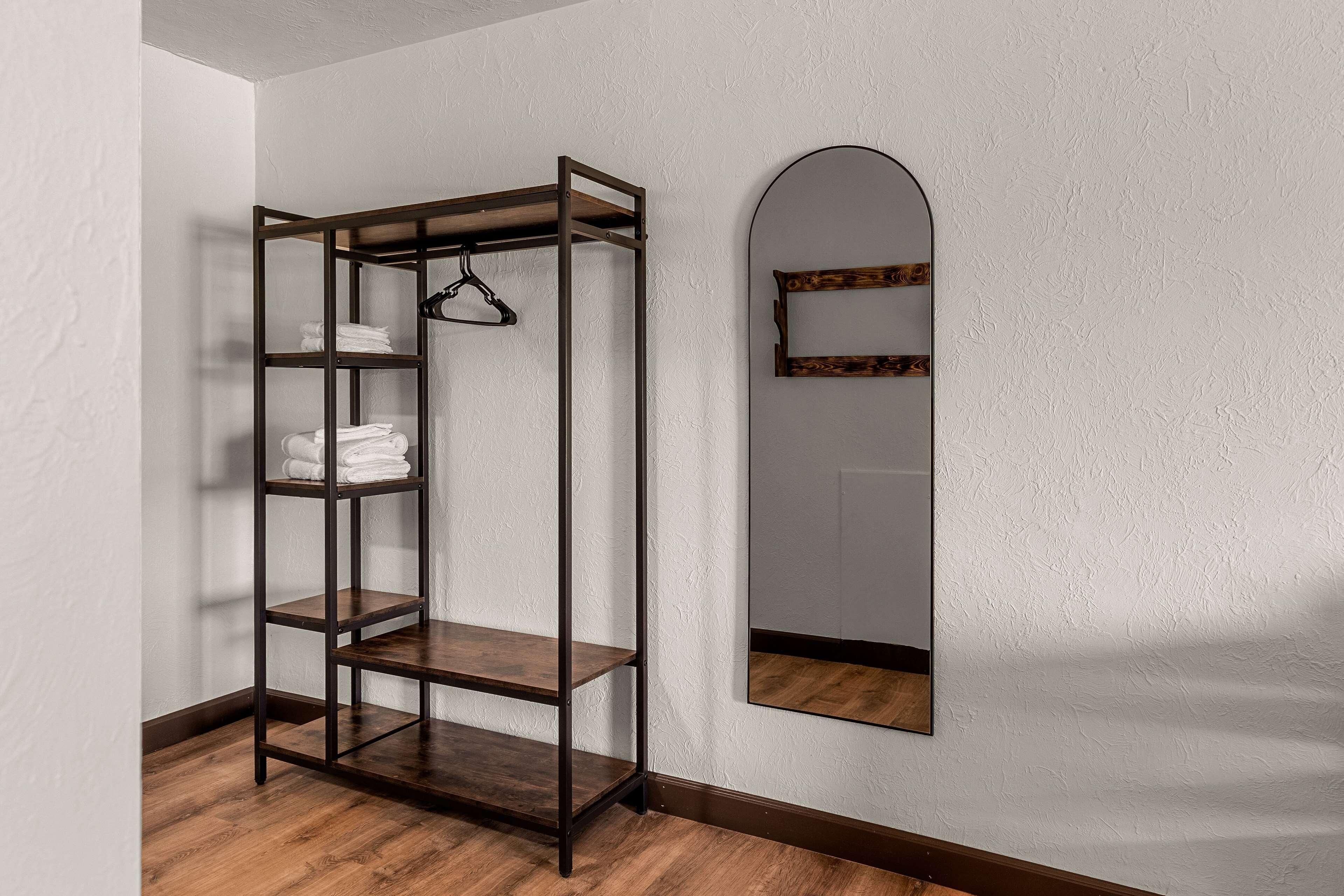 Una habitación de esquina con piso de madera, una estantería metálica con toallas y un espejo con marco de madera.