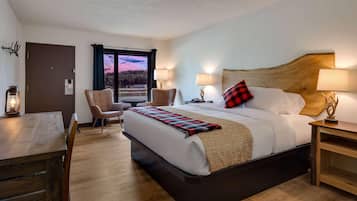 Una habitación de hotel con una cama grande, un escritorio y una silla. Se ve una ventana con vistas a árboles y al atardecer.