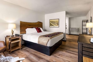 Een hotelkamer met een bed met houten hoofdeinde, een nachtkastje met een lamp, een ingelijst kunstwerk aan de muur, en een houten vloer.