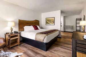 Quarto de hotel com cama de cabeceira em madeira, mesa de cabeceira com abajur, quadro emoldurado na parede e piso de madeira.