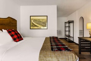 Ett hotellrum med en stor säng, en inramad tavla med en stadsbild på väggen, ett träfärgat sänggavel, en svart metallhylla och ett träbord med en lampa.