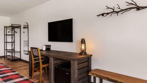A compact room with a wooden desk, a chair, a bench, a small refrigerator, a coat rack, and a wall-mounted TV. - Travelodge By Wyndham Emlenton (Emlenton)