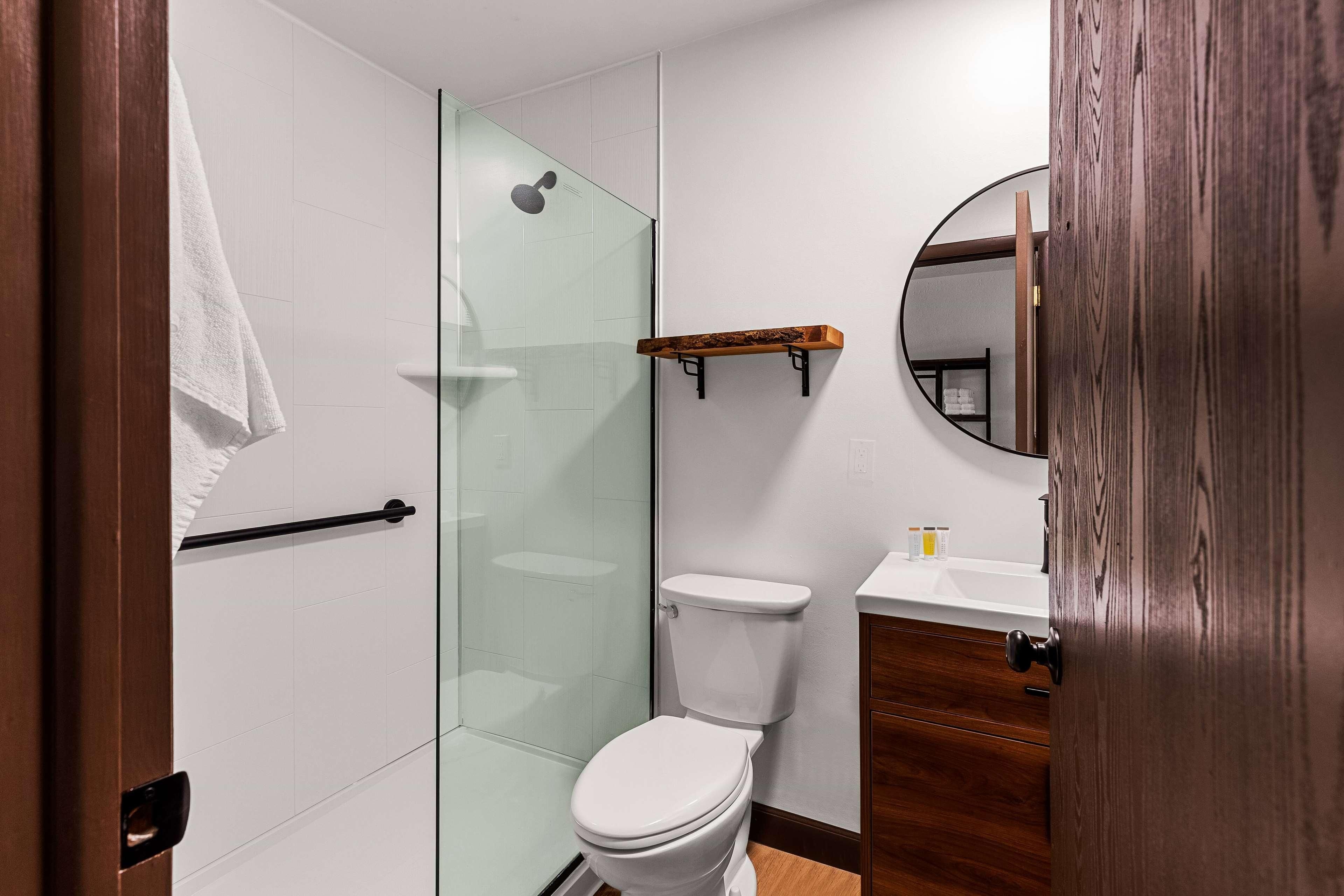 A modern bathroom with a glass shower enclosure, a white toilet, a wooden vanity, and a wooden door.