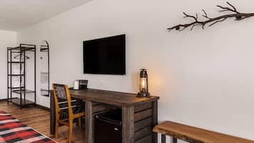 A compact room with a wooden desk, a chair, a bench, a small refrigerator, a coat rack, and a wall-mounted TV.
