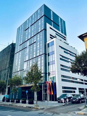 A modern multi-story hotel with a glass facade, a visible entrance, and a parking area with cars. - Elisa Hotel Tirana Affiliated By Melia (Tirana)
