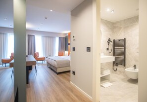 A modern hotel room with a wooden floor, a bed, a desk, and a chair. There is a bathroom with a towel rack, sink, and bidet. - Elisa Hotel Tirana Affiliated By Melia (Tirana)