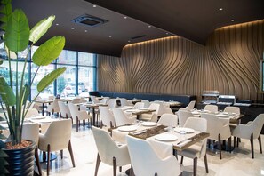 A modern restaurant with white chairs and tables, a large potted plant, and a wavy wall design. - Elisa Hotel Tirana Affiliated By Melia (Tirana)