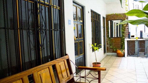 Um banco de madeira em um corredor com uma planta em vaso, uma porta com placa de número e uma parede de tijolos com um espelho.