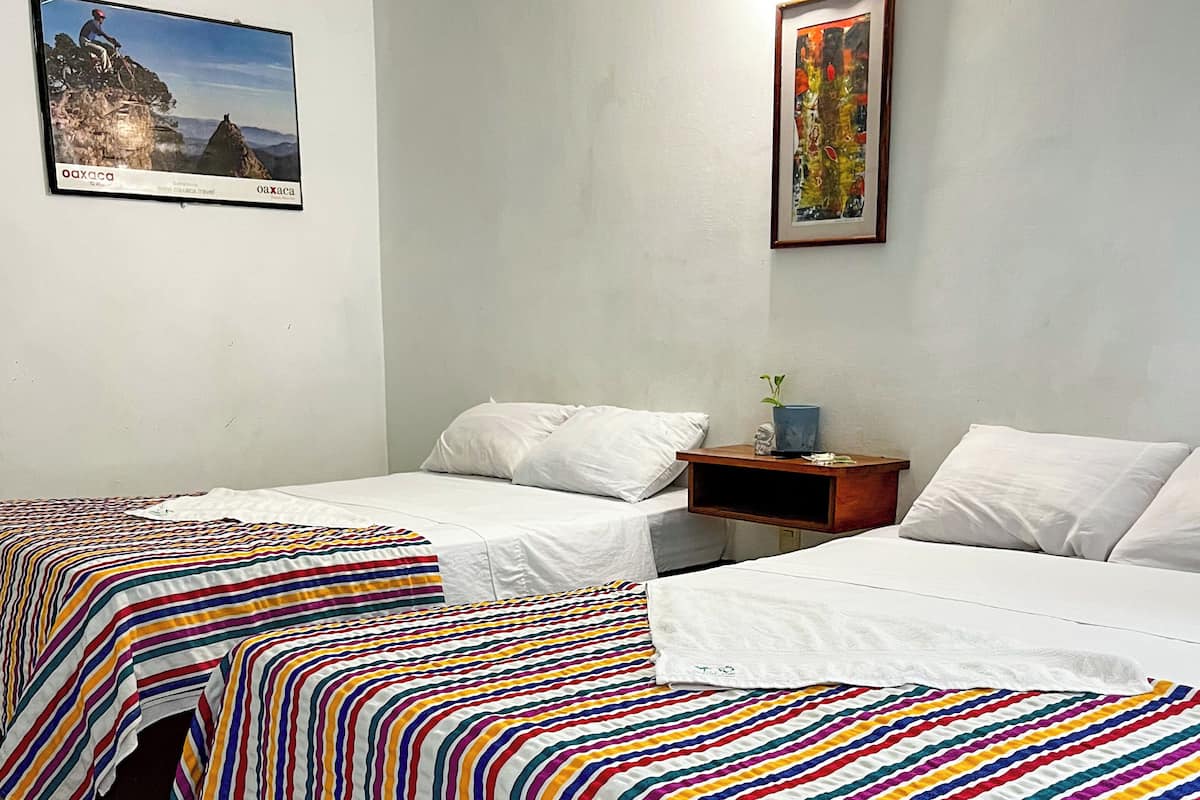 Une chambre d’hôtel avec deux lits, chacun recouvert d’une courtepointe à rayures colorées. On y trouve une petite table avec une plante et un tableau encadré accroché au mur.