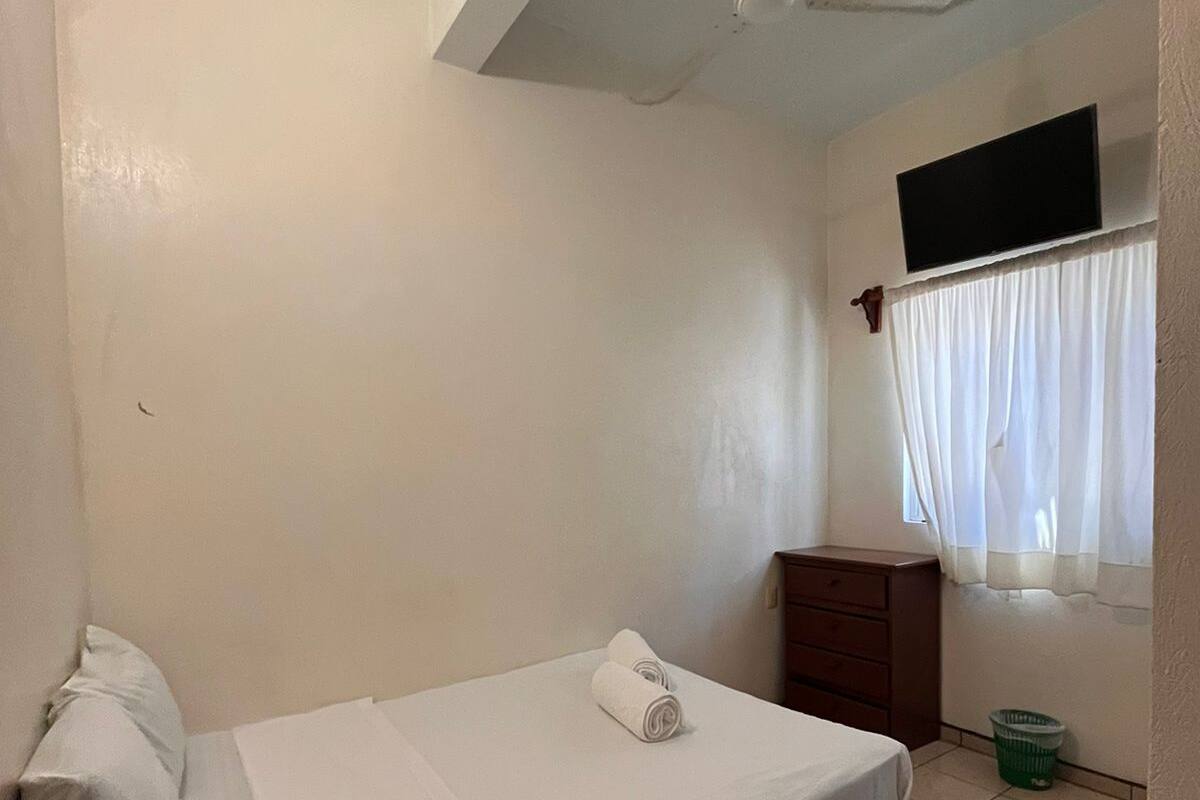 Une petite chambre propre, avec un lit simple, un ventilateur de plafond, un téléviseur fixé au mur et une fenêtre avec des rideaux.
