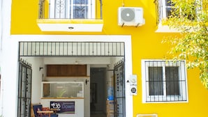 Um edifício amarelo com uma entrada ampla e aberta, uma pequena área de estar com mesa e cadeiras, e um bebedouro do lado de fora.