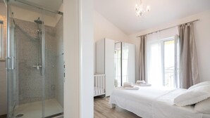 A modern bedroom with a glass-enclosed shower, a large bed with white linens, a wardrobe, and a chandelier. - Beautiful home in Grubine (Grubine)