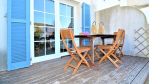 A wooden deck with a table and chairs, a blue door with shutters, and a view of the street. - Stunning home in Aigues-Mortes with WiFi (Aigues-Mortes)