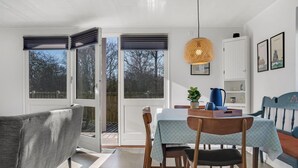 A dining area with a table set for two, a hanging light fixture, and a view through the open sliding doors to the outside.