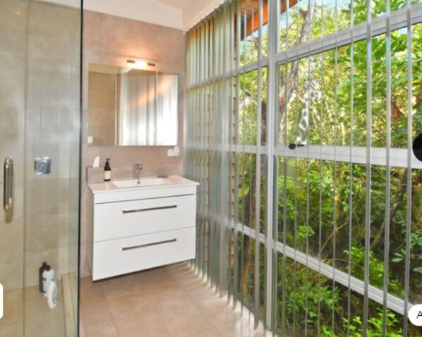 Ein modernes Badezimmer mit Duschkabine aus Glas, einem weißen Waschtisch mit Spiegel und Blick auf üppiges Grün durch ein großes Fenster.