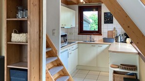 Cocina con armarios blancos, microondas y ventana. Se ve una escalera de madera que sube a otro nivel.