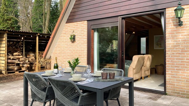 Un área de comedor al aire libre con una mesa dispuesta para cuatro personas, rodeada de sillas de mimbre, un muro de ladrillo y una puerta de cristal que da a un espacio interior.