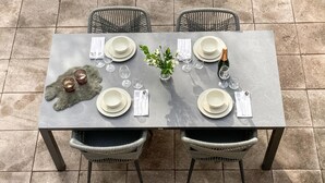 Mesa de comedor preparada para cuatro personas, con platos, vasos y cubiertos. Hay dos sillas de mimbre y una planta pequeña sobre la mesa.