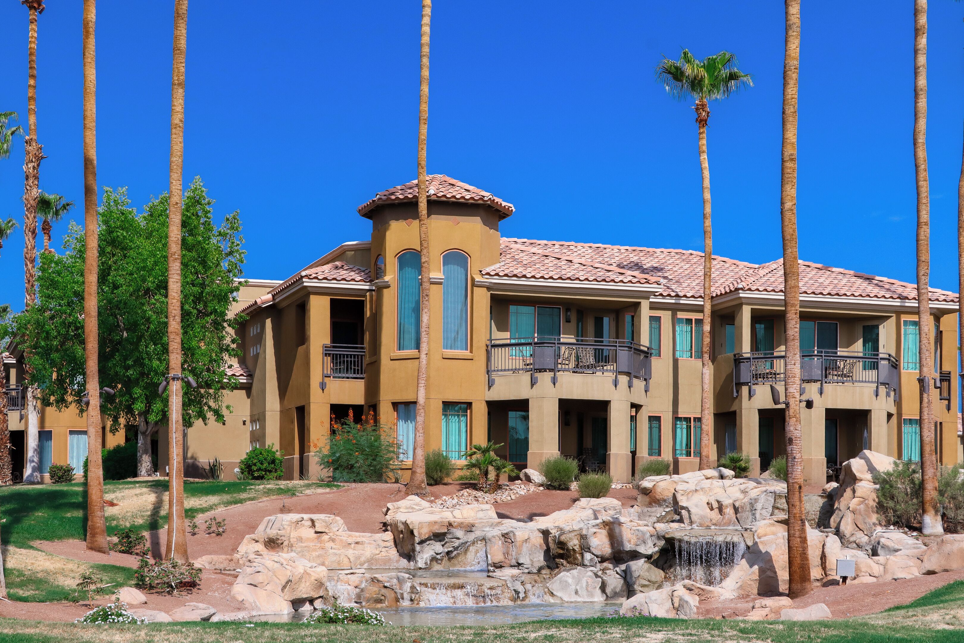 A multi-story hotel with a Mediterranean architectural style, featuring balconies and a landscaped garden with palm trees and a rock waterfall.
