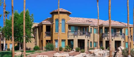 A multi-story hotel with a Mediterranean architectural style, featuring balconies and a landscaped garden with palm trees and a rock waterfall.