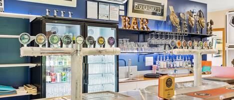 A bar area with a beer tap display, a refrigerator stocked with beverages, and a shelf with glasses.