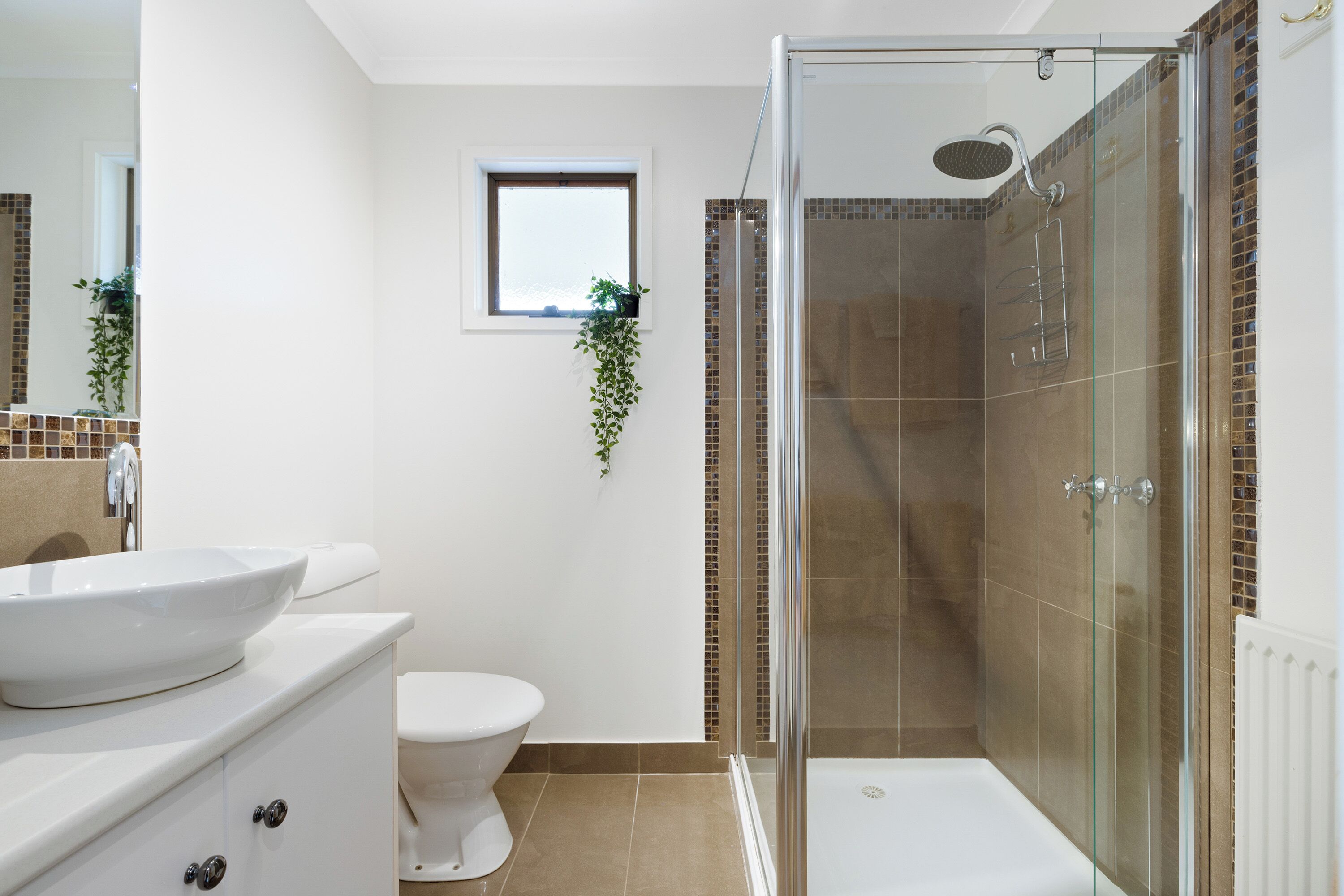 A bathroom with a glass-enclosed shower, a white toilet, a large white sink, and a small window with a plant.