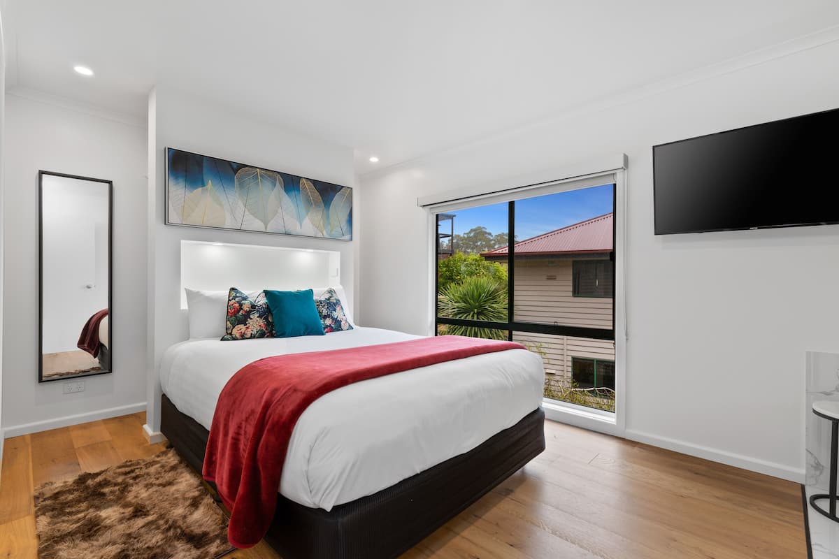 A modern bedroom with a large bed, a flat-screen TV, a painting, and a view of the outside.