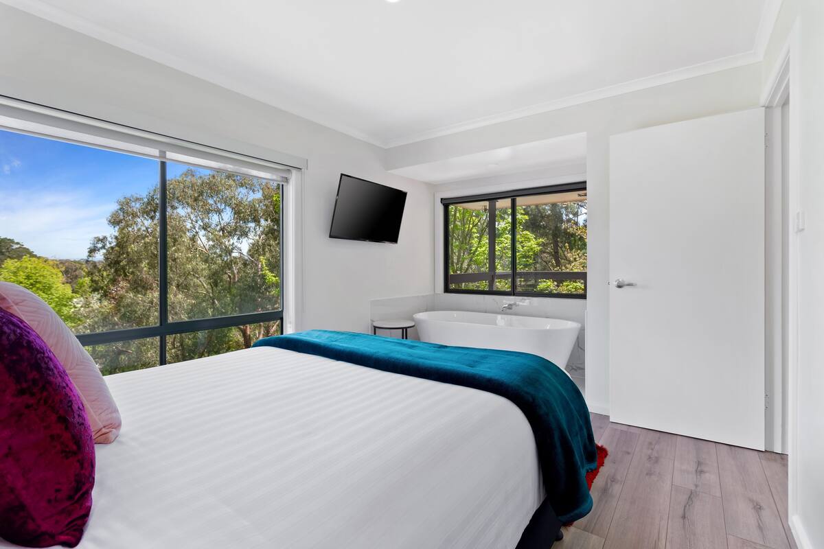 A modern bedroom with a large bed, a flat-screen TV, a bathtub, and a view of trees through large windows.