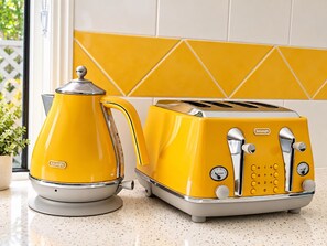 Une bouilloire électrique et un grille-pain jaunes de la marque DeLonghi sont posés sur un plan de travail de cuisine, devant un mur recouvert de carreaux.