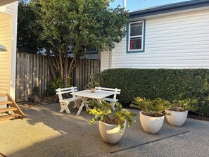 Une terrasse avec une table blanche et des bancs, entourée de plantes en pot et d’une clôture en bois.