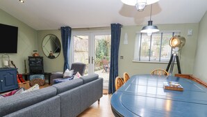 En stue med en grå sofa, et spisebord med stole, et TV ophængt på væggen og et vindue med gardiner.