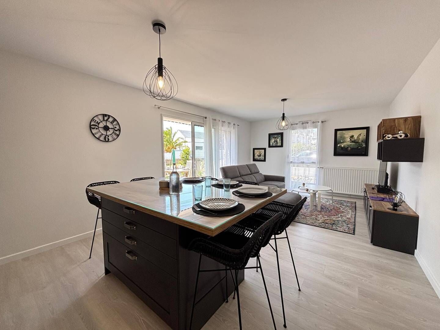 A modern kitchen with a central island, dining area with chairs, a sofa, and a coffee table. There are wall-mounted clocks and framed pictures.