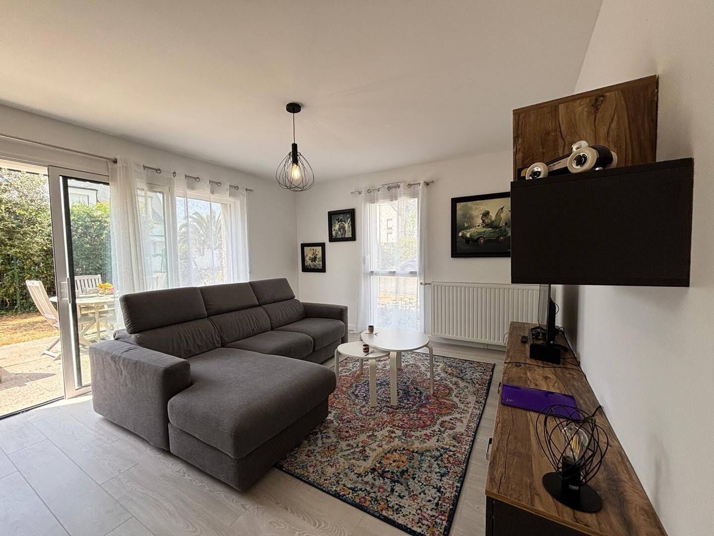 A modern living room with a grey sofa, a small round table, and a colorful rug. There are two windows with curtains, a ceiling light, and a flat-screen TV mounted on the wall.