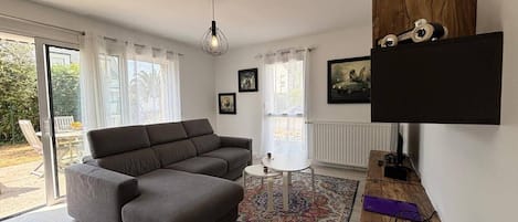 Una sala de estar moderna con un sofá gris, una mesita redonda pequeña y un tapiz colorido. Hay dos ventanas con cortinas, una lámpara de techo y un televisor de pantalla plana montado en la pared.