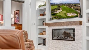 A living room with a fireplace, a mounted TV displaying a golf course, and a patterned armchair. - Scottsdale Grayhawk entire house with Private Pool, Sunroom, Office (Scottsdale)