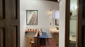 A narrow hallway with a wooden bench, a small shelf with potted plants, a framed picture on the wall, and a glimpse of a bathroom with a sink. - PiYo (San Cristóbal de las Casas)