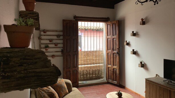 A cozy room with a wooden ceiling, a wooden door leading outside, a small table with a plant, and a television on a wooden cabinet. - PiYo (San Cristóbal de las Casas)