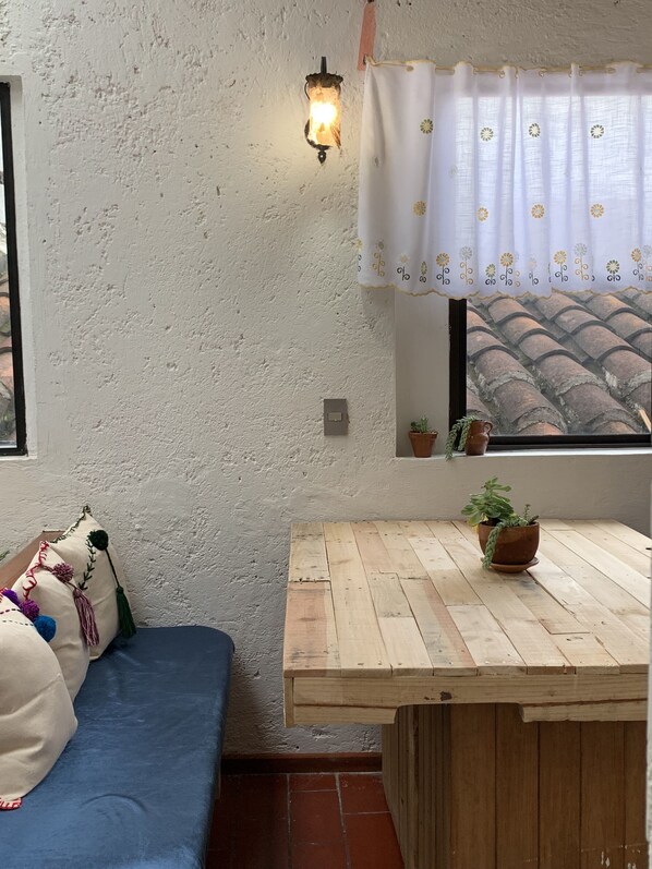 Habitación acogedora con sofá azul, una mesa de madera, una lámpara de pared y una ventana con vista a un tejado de baldosas.