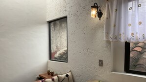 A cozy room with a wooden table, a potted plant, a cushioned bench, a wall-mounted light fixture, and a window with a curtain. - PiYo (San Cristóbal de las Casas)