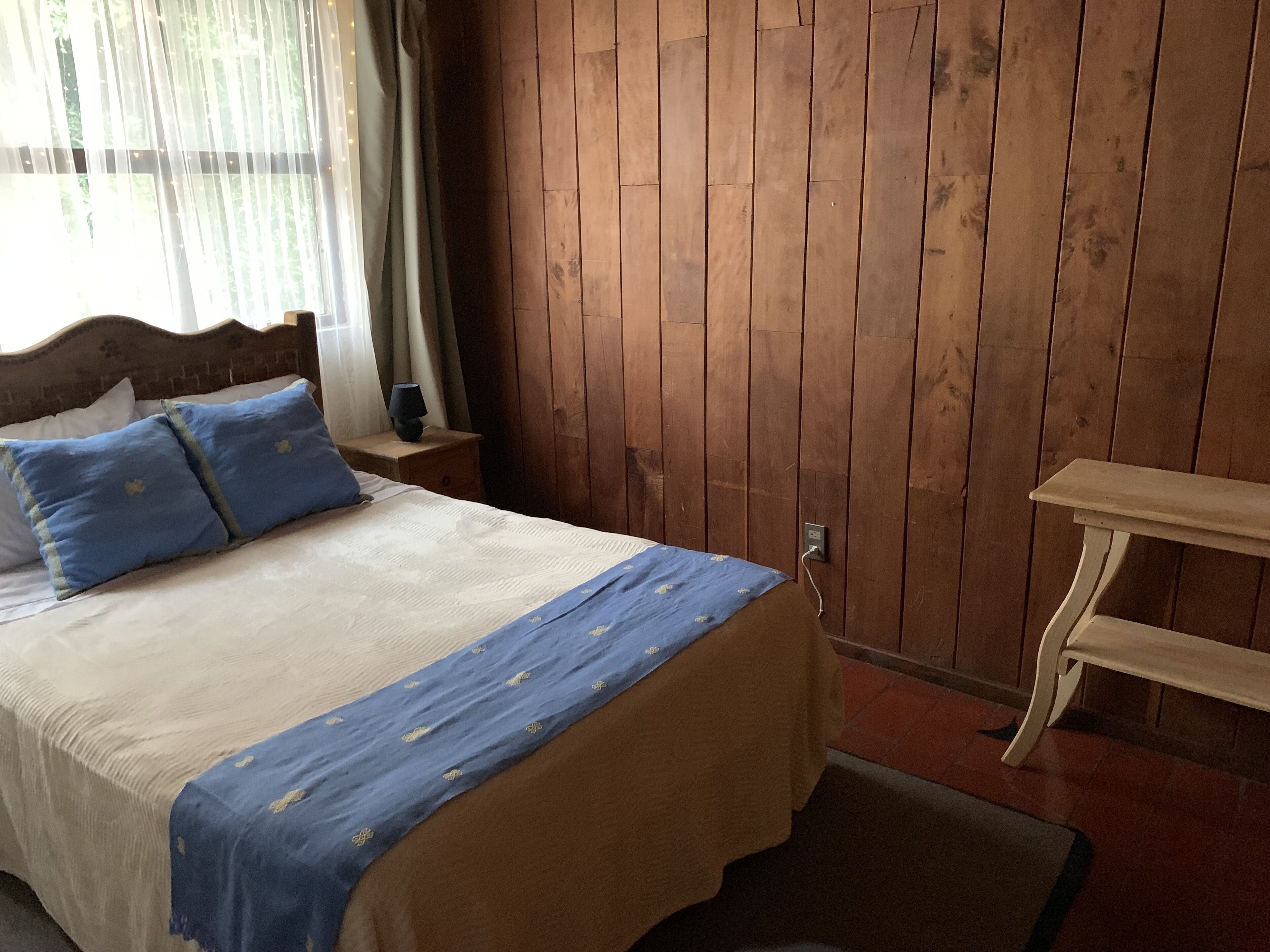 Une chambre à coucher avec une tête de lit en bois, un lit recouvert d’une couvre-lit bleue et beige, une table de chevet avec une lampe et un banc en bois.