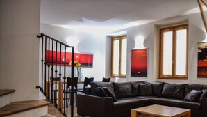 Ein Wohnzimmer mit einer schwarzen Ledersofa, einem Holztisch und einem Essbereich mit Tisch und Stühlen. Zwei Fenster mit Vorhängen sind zu sehen, sowie eine Treppe mit schwarzer Geländer.