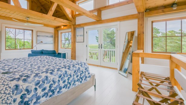Une chambre spacieuse dotée d’un grand lit, de poutres en bois et de nombreuses fenêtres offrant une vue sur l’extérieur.