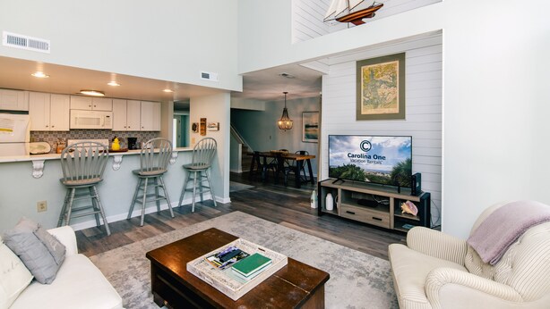 Een moderne keuken met barstoelen, een eethoek met een televisie waarop 'Carolina One Vacation Rentals' staat, en een woonkamer met een salontafel.