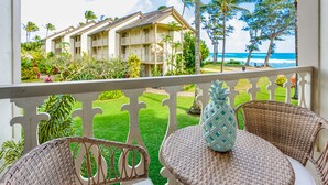 A balcony with wicker furniture and a pineapple decoration, overlooking a hotel with a lawn and palm trees, and a view of the ocean. - Drift Away |Studio Oceanview with AC (Kapaa)