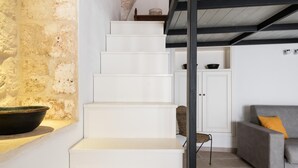 Eine Treppe führt zu einem Erkerbereich mit einer rustikalen Steinwand, einem grauen Sofa und einem Korbsessel.