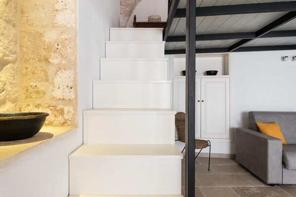 A staircase leading to a loft area with a rustic stone wall, a grey sofa, and a wicker chair.