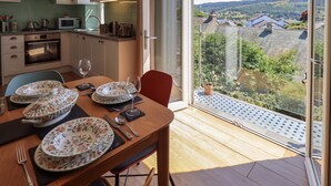 Ein Essbereich mit Tisch für vier Personen, eine Küche im Hintergrund und Blick auf Häuser durch ein großes Fenster.