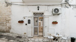 A white building with a stone ground floor, a white door with a fan above it, and a small outdoor seating area with a table and chairs.