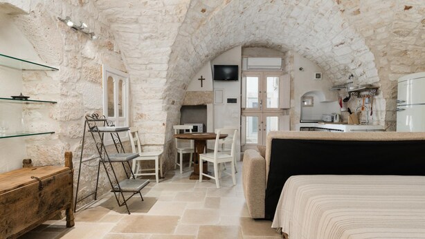 A cozy, rustic room with a stone ceiling, a bed, a dining table with chairs, a TV, and a kitchenette.