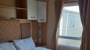 A neatly made bed with white bedding, a headboard with storage compartments, and a window with curtains. - Northfield 198, Skipsea Sands (Driffield)